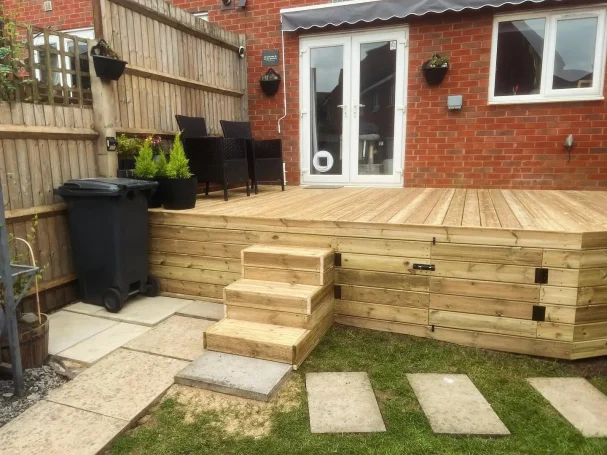 Wooden deck with steps, chairs, and a bin in a garden area.