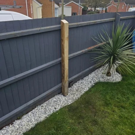 A wooden fence with a concrete base, bordered by gravel and a small palm plant.