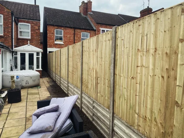 A wooden fence separates two back gardens with patio furniture in the foreground.