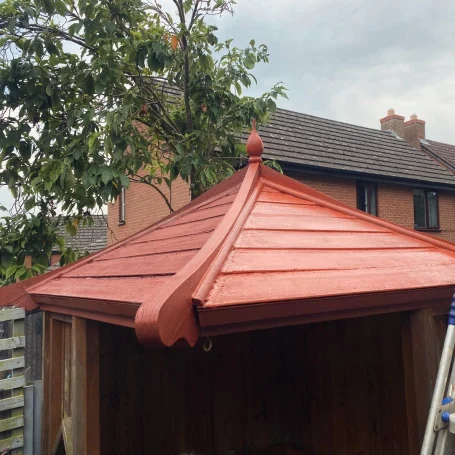 Red-tiled roof of a gazebo with decorative peak, surrounded by buildings and trees.