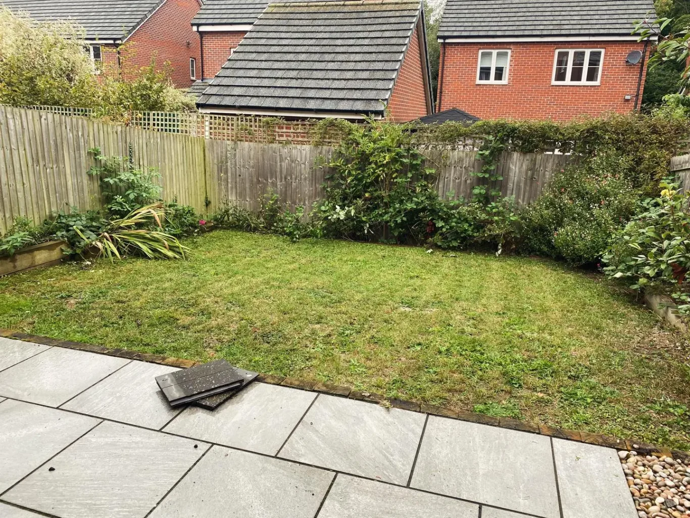 A manicured garden with a paved area and green grass, bordered by shrubs and houses.