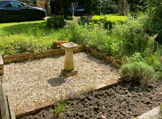 A gravel garden with a stone birdbath surrounded by greenery and flowers.